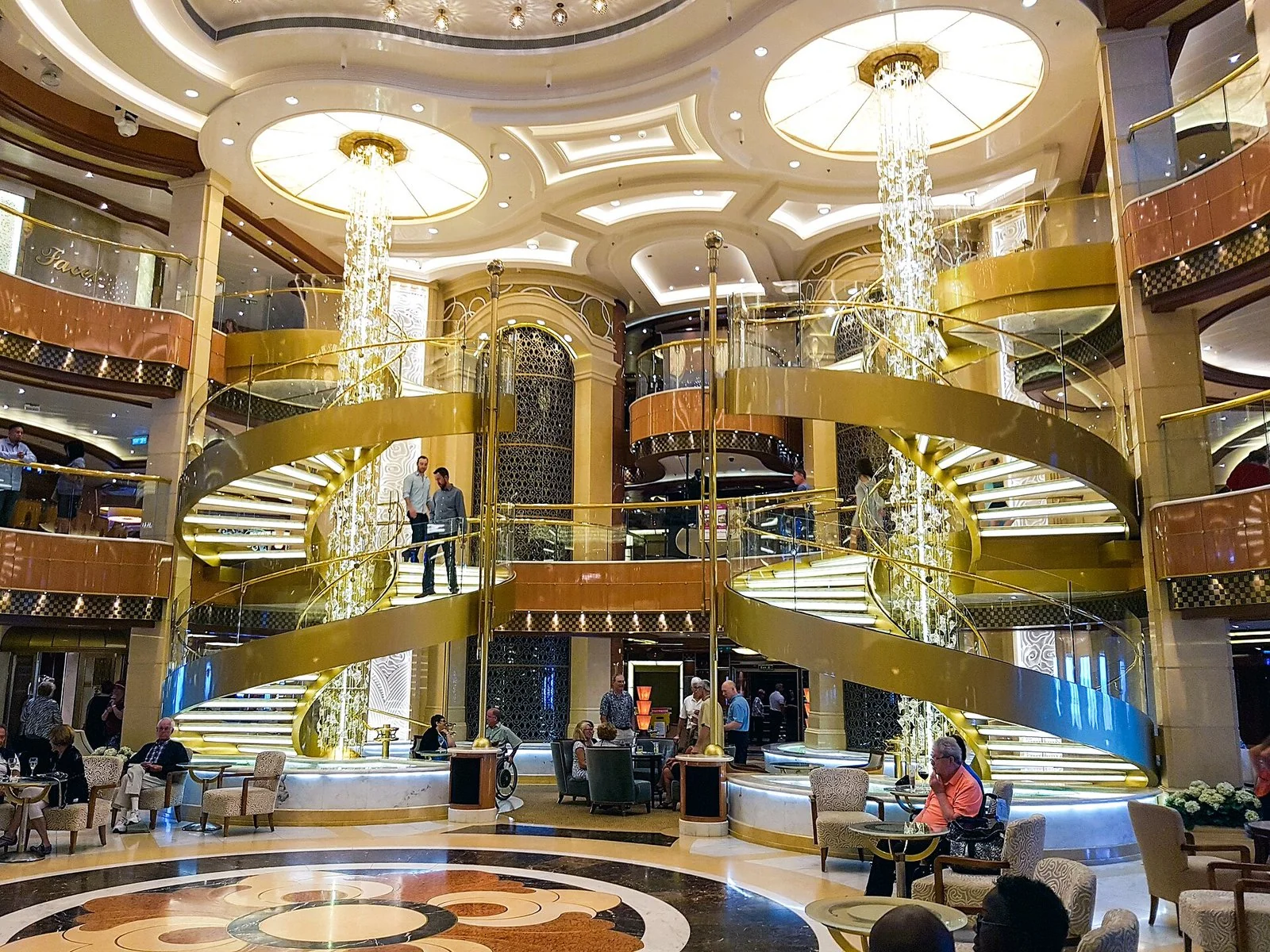 客船の吹き抜けの広場。金色のらせん階段が2つと、天井から下がる長い照明。下にソファの席が並ぶ。