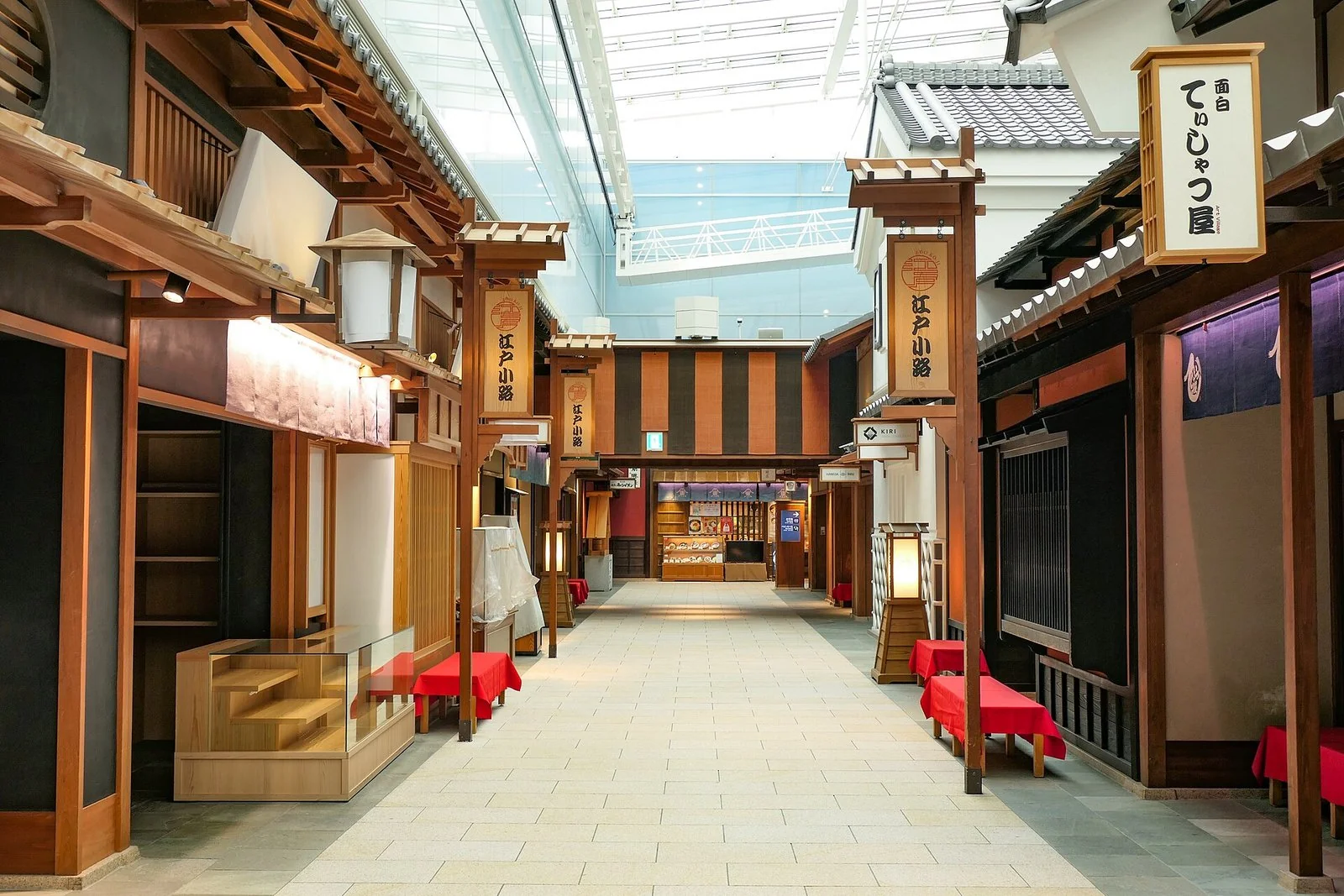 ガラスの天井の下に、木の柱と瓦の屋根の店が両側に並ぶ通り。店の前に赤い布の腰掛けがある。