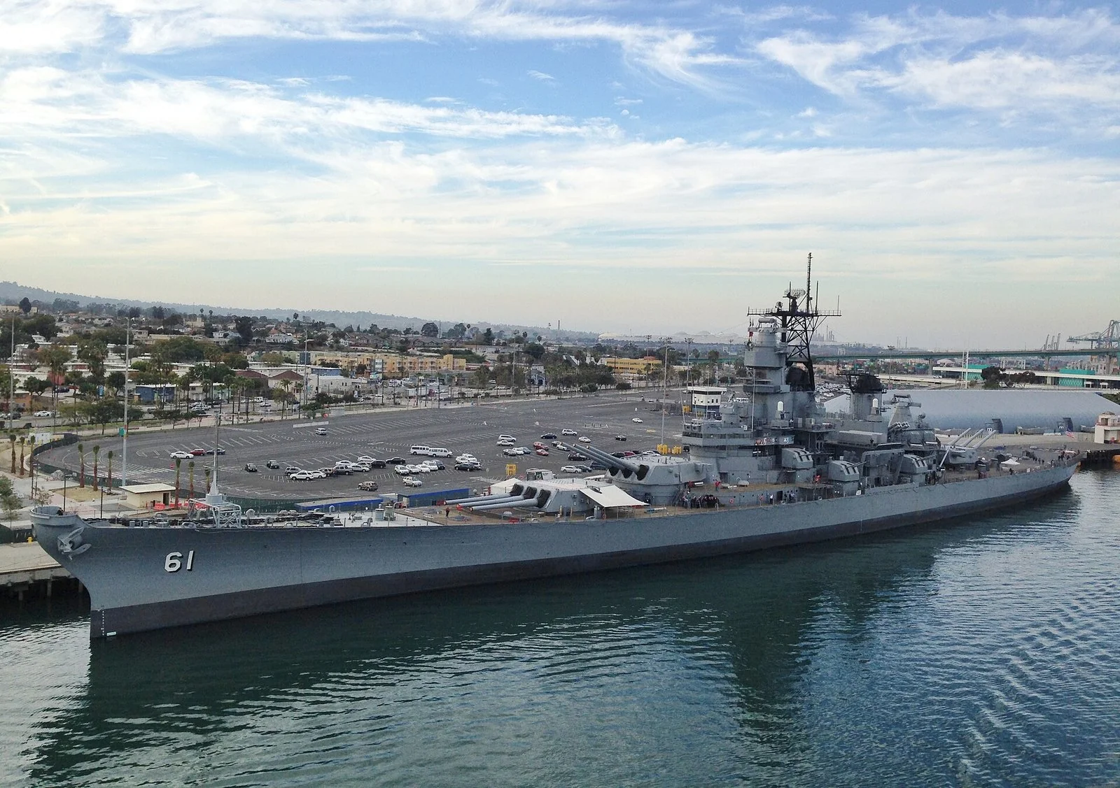 青い空の下、岸壁に泊まる灰色の大きな戦艦。船首に61の番号、甲板に大きな砲塔が並び、奥に駐車場と町が見える。