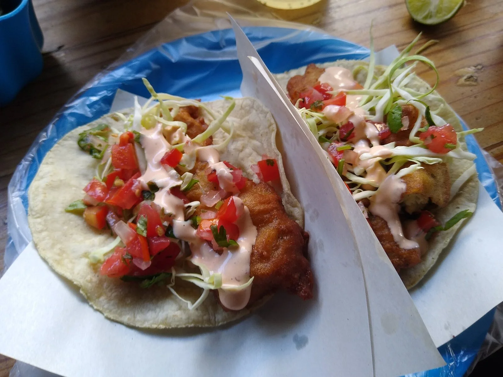 紙を敷いた青い皿に、揚げた魚とキャベツ、トマトのサルサ、ピンクのソースをのせたタコスが2つ。