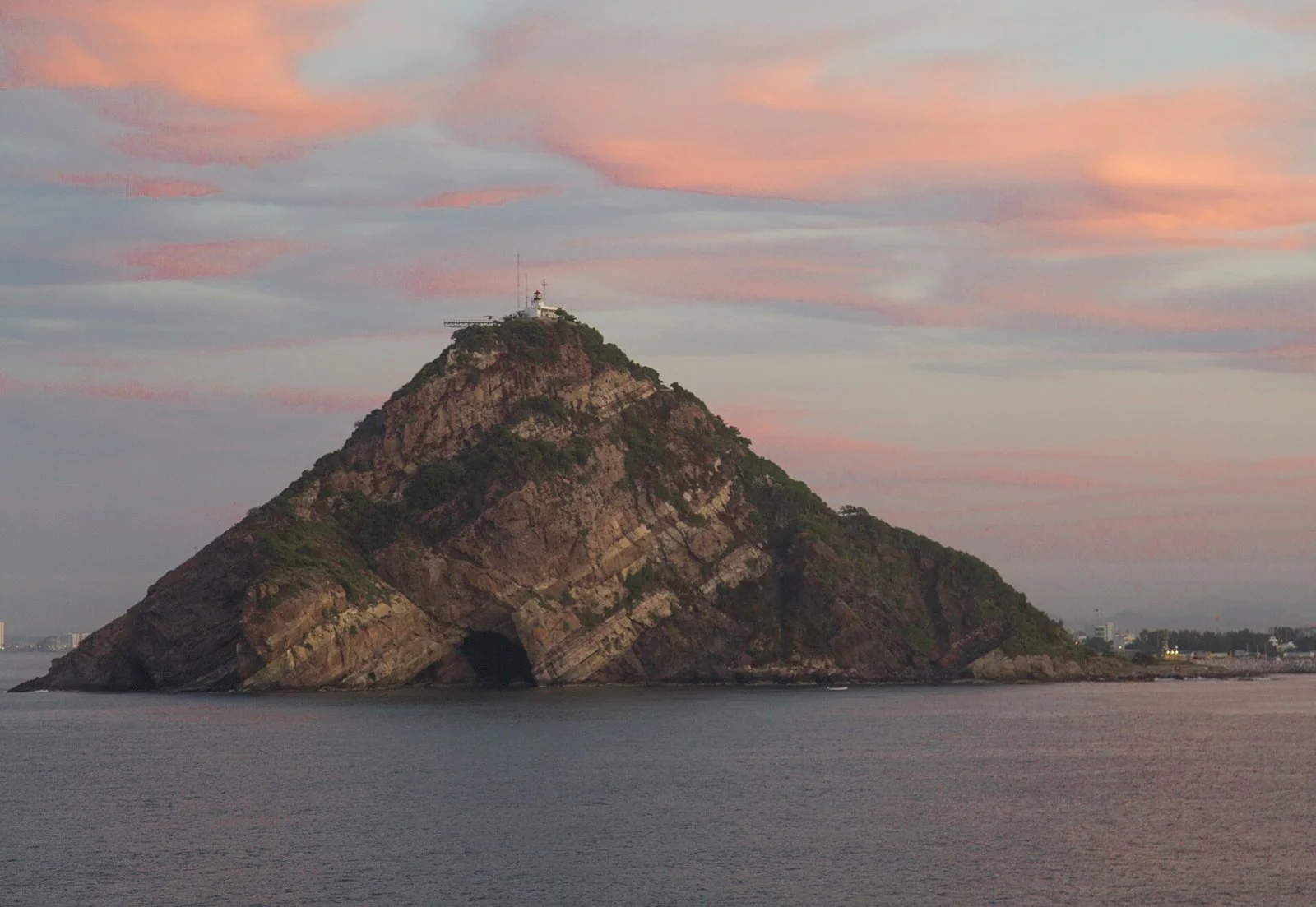 朝焼けの雲の下、海から三角に立ち上がる岩山と、その頂の白い灯台。右の奥に町の建物が見える。