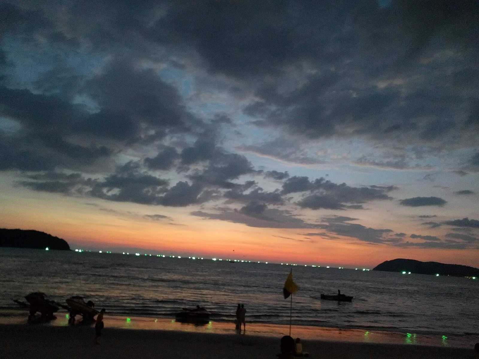 日の入りのあとの浜。雲の切れ間に夕焼けが残り、沖の島の影と水平線に船の明かりが並ぶ。