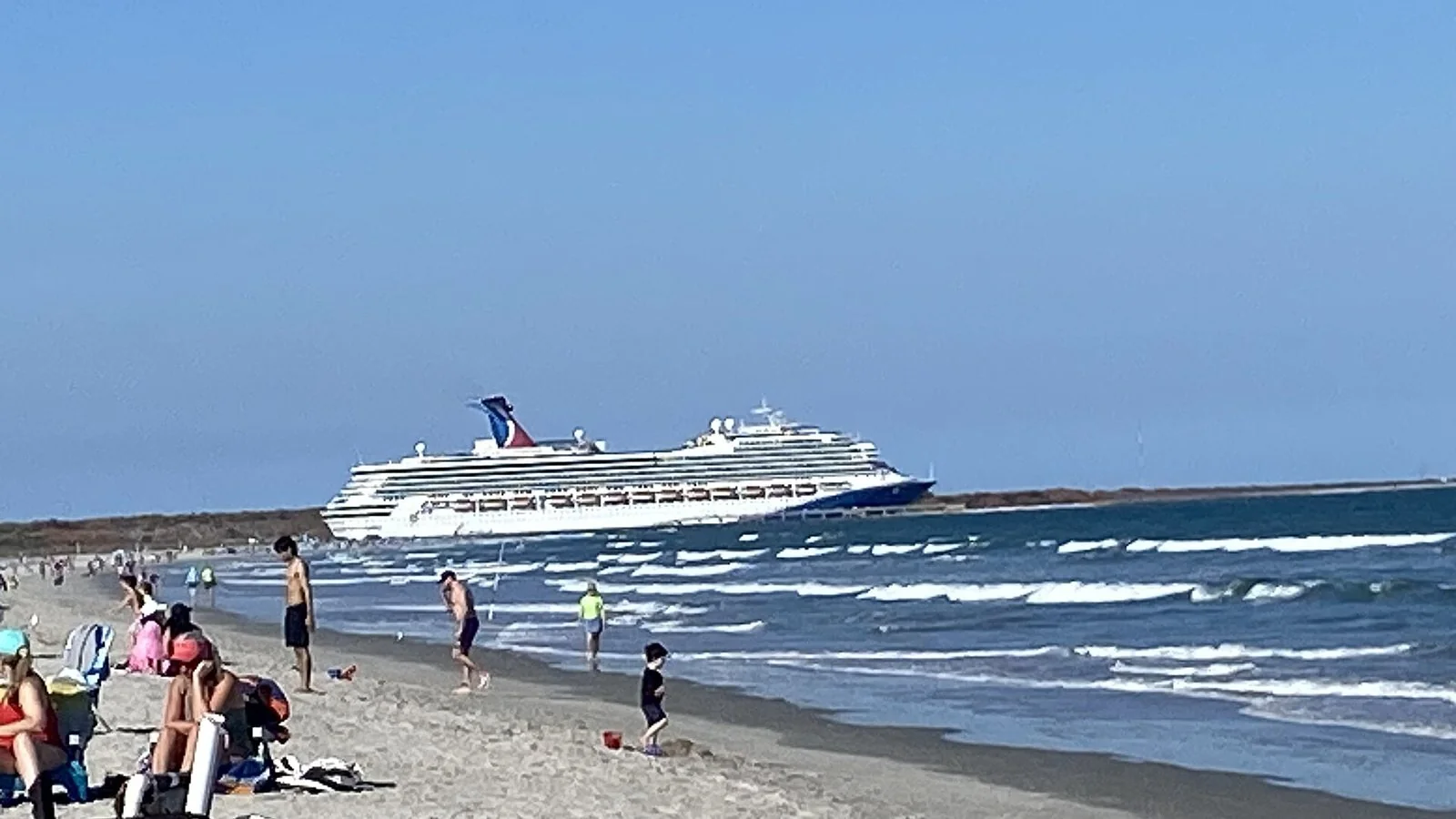 晴れた空の下、波の寄せる砂浜の沖を、青い煙突の白い大型客船が横切っていく。浜では人が水際を歩いている。