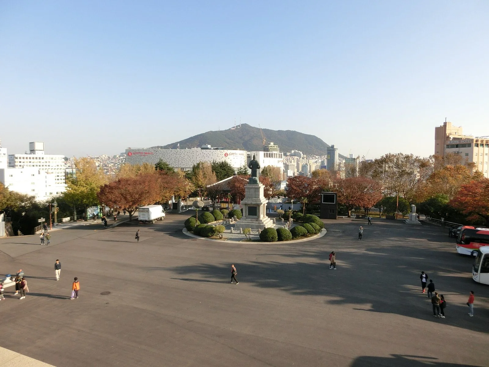 広い石畳の広場の中央に像が立ち、まわりに紅葉した木と歩く人の姿がある。