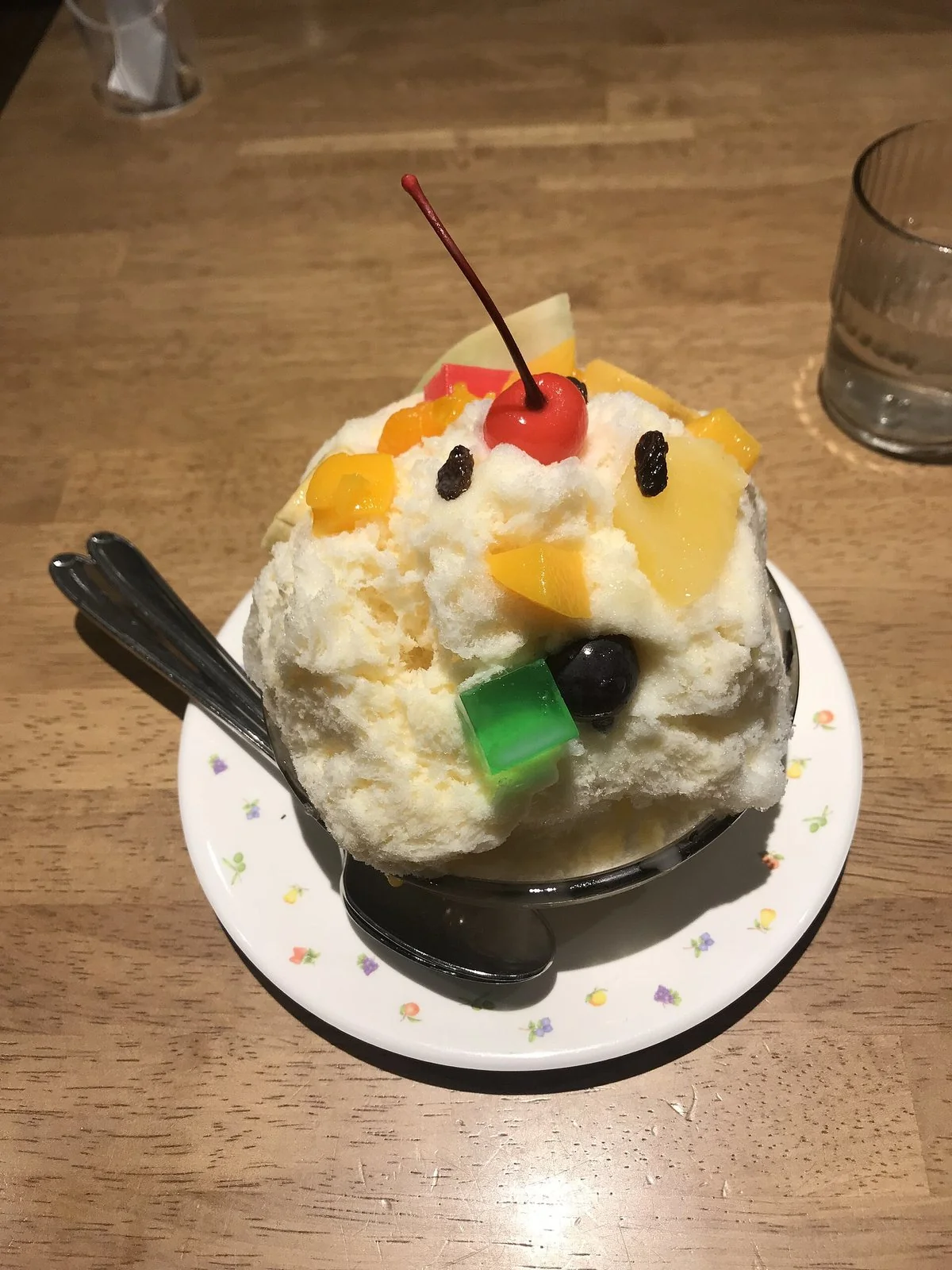 皿にのった山盛りのかき氷。上にさくらんぼ、レーズン、黄色い果物、緑のゼリーがのり、顔のように見える。