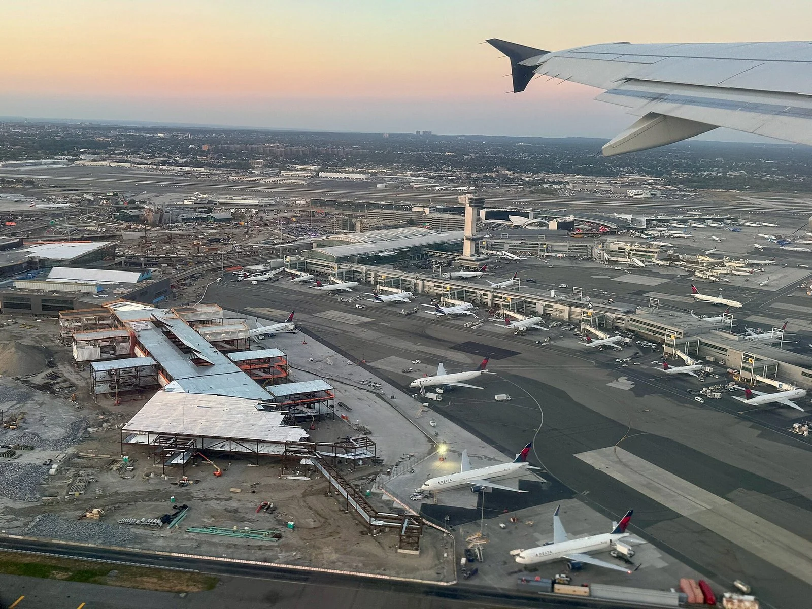 夕方の空の下、飛行機の翼の先に空港が広がり、工事中の建物の横に旅客機が並ぶ。