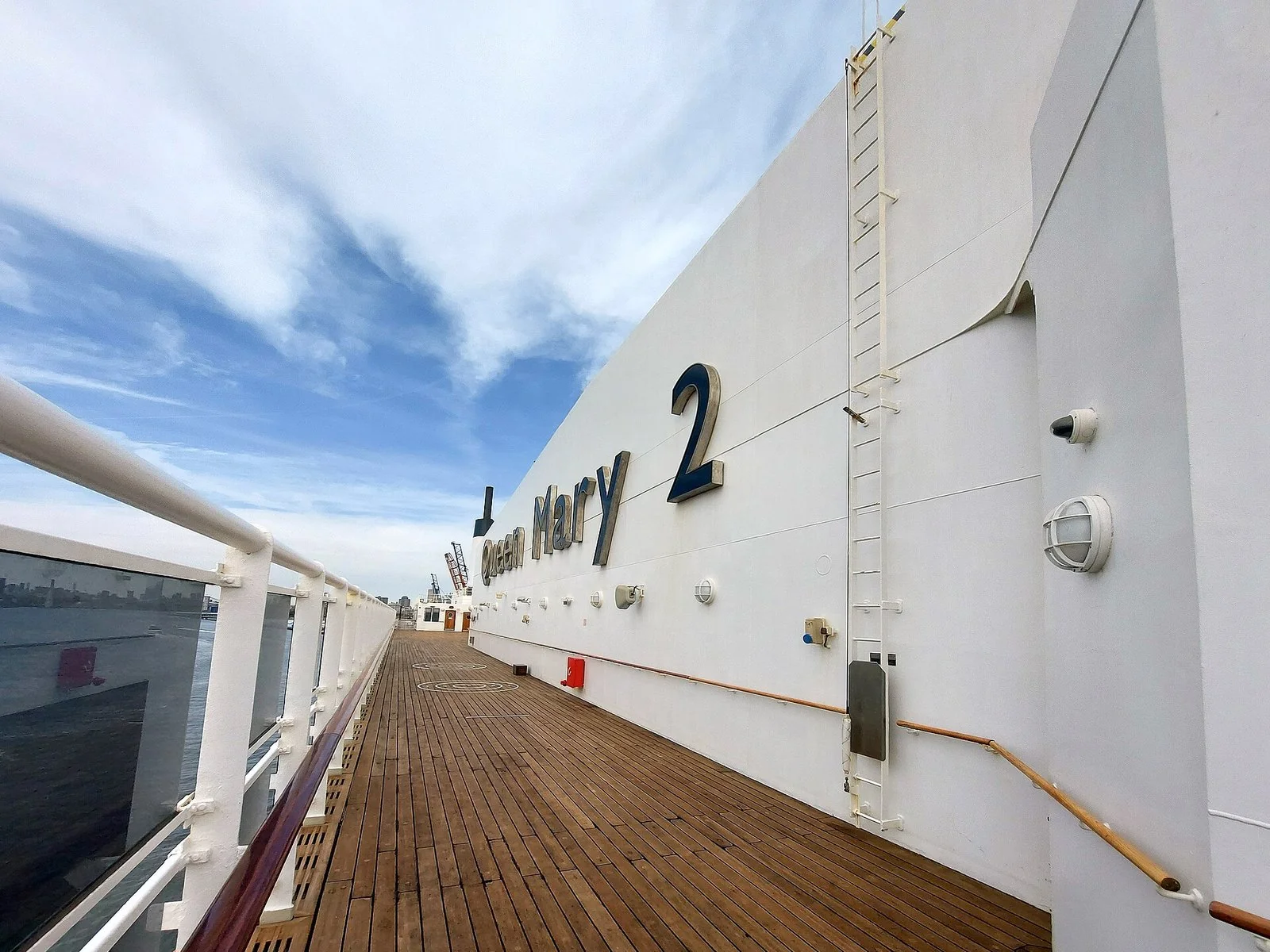 青空と雲の下、木の甲板がまっすぐ奥へ延び、右の白い壁に「Queen Mary 2」の大きな文字がある。