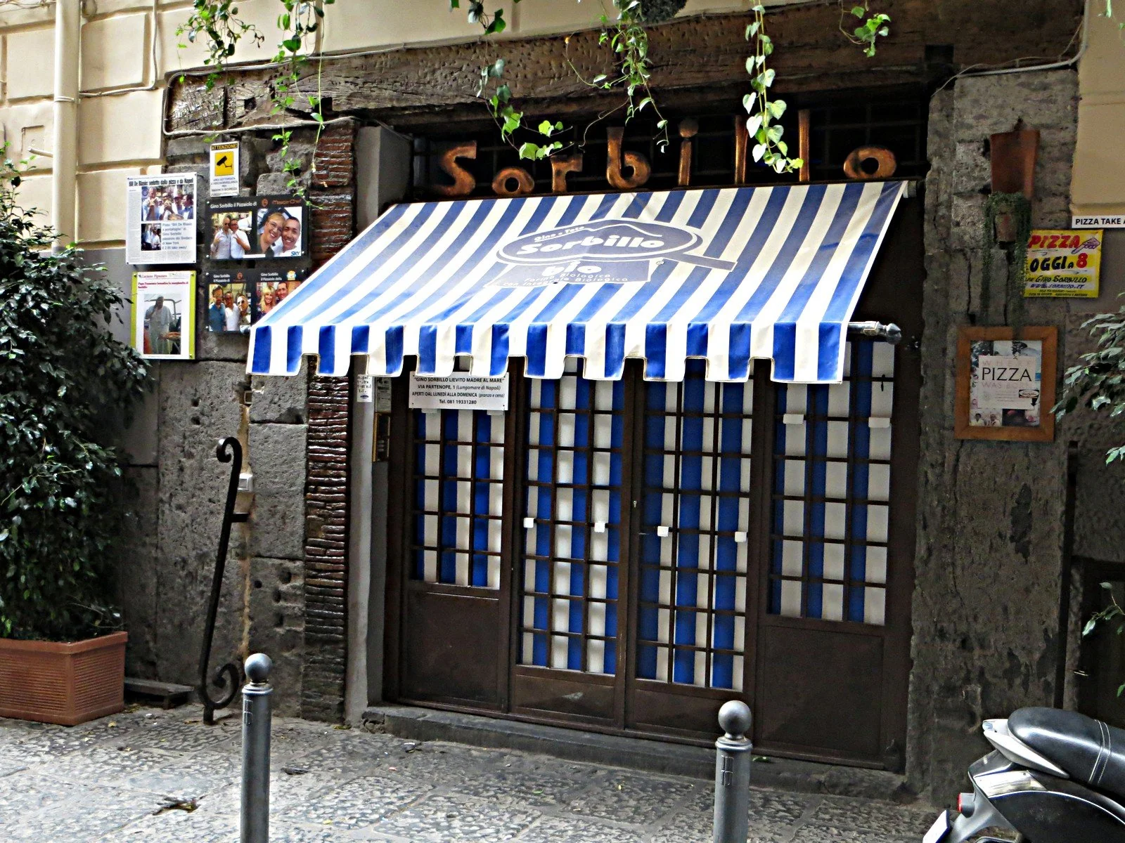 石の壁の建物に、青と白の縞の日よけと店の名前の文字。閉じた格子の扉と、壁に貼られた記事。